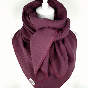 Puede incluir: Bufanda de color ciruela con un aspecto texturizado y arrugado. La bufanda está drapeada alrededor de una cabeza de maniquí blanca y tiene un ribete rojo oscuro en contraste. La tela parece suave y ligera, adecuada para climas más frescos.