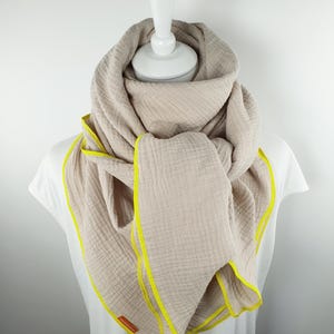 Puede incluir: Una bufanda de algod&oacute;n beige con un borde amarillo. La bufanda est&aacute; doblada y drapeada sobre un maniqu&iacute; blanco.