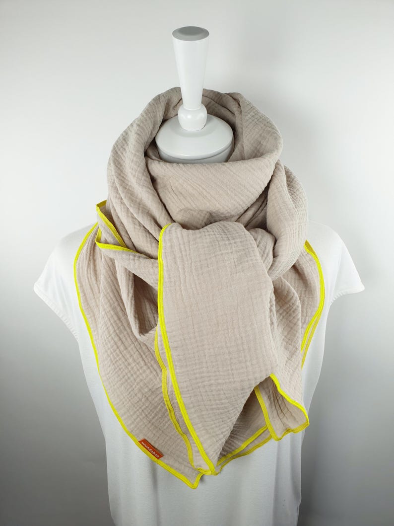 Puede incluir: Una bufanda de algod&oacute;n beige con un borde amarillo. La bufanda est&aacute; doblada y drapeada sobre un maniqu&iacute; blanco.