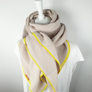 Puede incluir: Una bufanda beige con ribete amarillo. La bufanda est&aacute; doblada y drapeada alrededor de un maniqu&iacute; blanco.