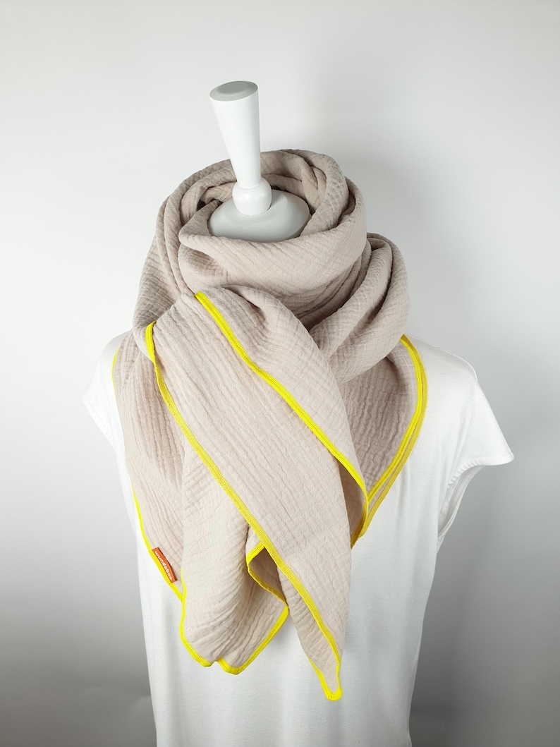 Puede incluir: Una bufanda beige con ribete amarillo. La bufanda est&aacute; doblada y drapeada alrededor de un maniqu&iacute; blanco.