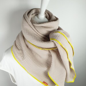 Puede incluir: Una bufanda de algod&oacute;n beige con ribete amarillo. La bufanda est&aacute; doblada y drapeada sobre un maniqu&iacute; blanco. El nombre de la marca "woodstock" es visible en la bufanda.