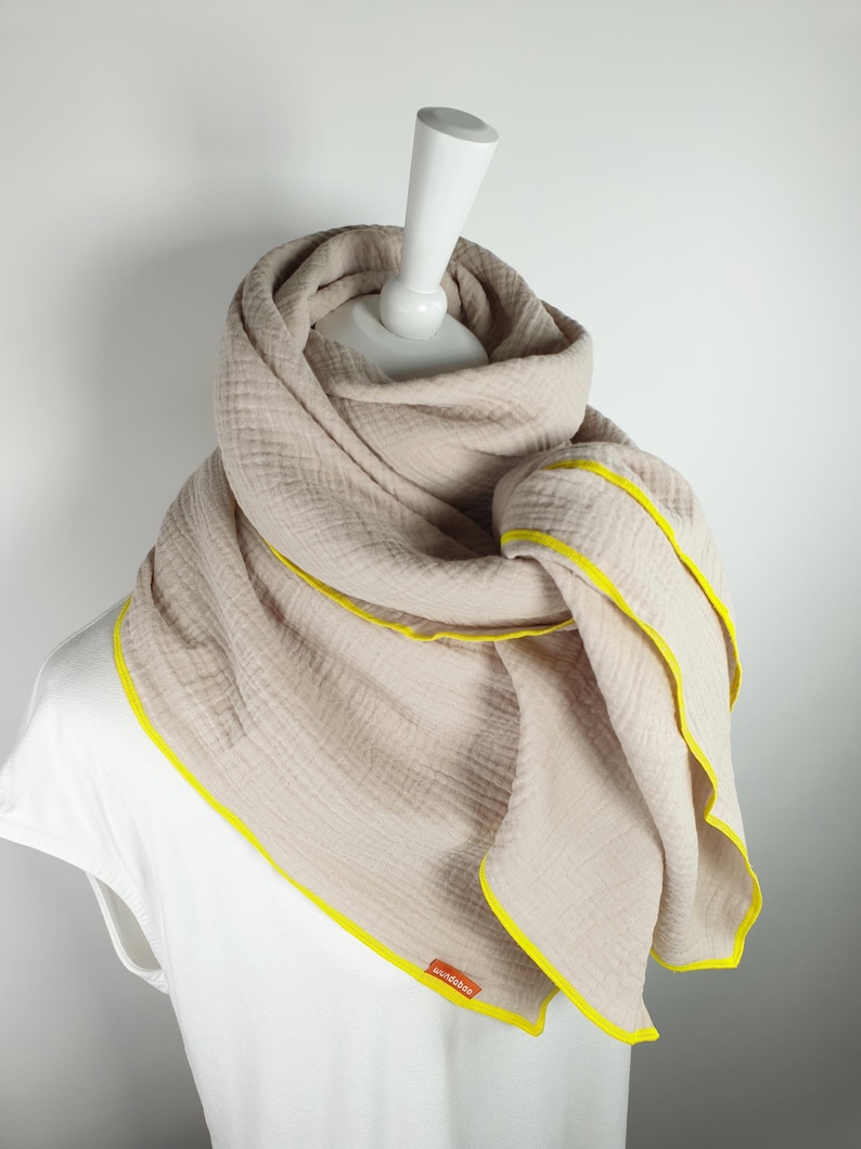 Puede incluir: Una bufanda de algod&oacute;n beige con ribete amarillo. La bufanda est&aacute; doblada y drapeada sobre un maniqu&iacute; blanco. El nombre de la marca "woodstock" es visible en la bufanda.