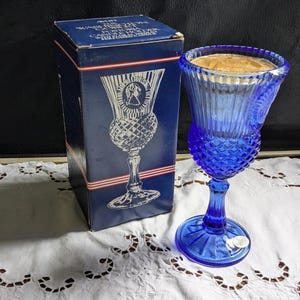 May include: A blue glass candle holder with a diamond pattern and a gold medallion with a woman's profile. The candle holder is in its original box, which is blue with red and white stripes.