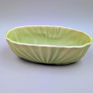 May include: A light green ceramic oval-shaped bowl with a scalloped design. The bowl has a glossy finish and a slightly off-white rim. The ridges radiate from the center, creating a textured appearance. This decorative bowl is suitable for serving or display.