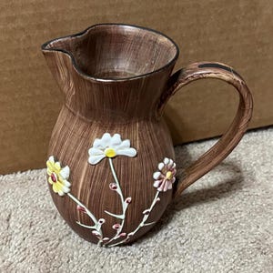 May include: A brown ceramic pitcher with a wooden-grain design and three white, yellow, and pink flowers with green stems.