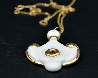 May include: A gold chain necklace featuring a white ceramic pendant with a central gold ring, gold-toned edges, and two small gold accents on the sides. The pendant has a smooth, glossy finish and is suspended from a delicate gold-colored metal chain.