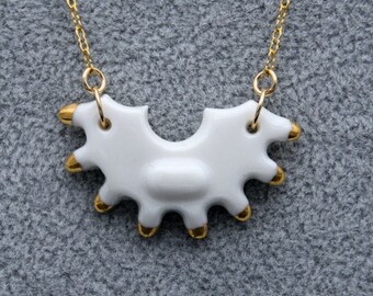 May include: A white ceramic pendant with a curved, multi-pronged shape and gold-painted tips, suspended from a delicate gold-tone metal chain. The pendant features a smooth, glossy finish and is attached to the chain by two gold jump rings.