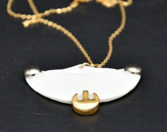 May include: A necklace featuring a white, semi-circular ceramic pendant suspended from a thin gold-tone chain. The pendant is accented with two small silver-tone metal studs at the top corners and a gold-tone metal piece at the bottom center.