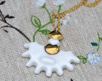 May include: A white ceramic pendant with a fan-like shape and gold-colored metallic accents at the top, suspended from a thin gold-colored chain. The pendant features a smooth, glossy finish and a series of rounded, protruding points along the bottom edge.