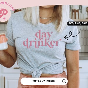 Könnte beinhalten: Ein graues T-Shirt mit dem Text "day drinker" in rosa Schrift. Das Shirt ist in der Taille gebunden und das Model hält eine rosa-weiße Tasse. Das Bild zeigt auch ein Schild mit der Aufschrift "Pour some sugar on me - coffee".