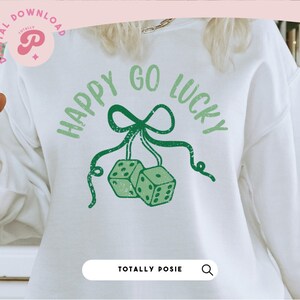 Puede incluir: Sudadera blanca con texto verde que dice "Happy Go Lucky" con un lazo verde y dos dados colgando del lazo.