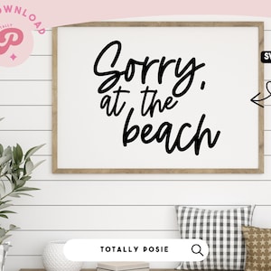 Peut inclure: Une affiche encadrée avec le texte "Désolé, à la plage" en écriture cursive noire. L'affiche est accrochée à un mur blanc en lambris. L'image comprend également une plante verte, deux coussins et une surface en bois.