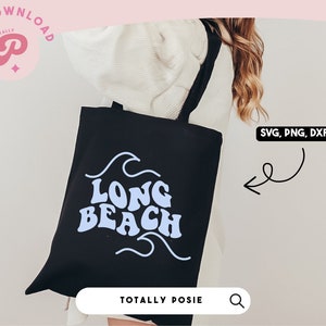 Puede incluir: Una bolsa de tela negra con un diseño azul y blanco que dice "Long Beach" en una fuente retro. La bolsa está siendo sostenida por una persona que lleva un suéter blanco.