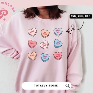Könnte beinhalten: Ein rosa Sweatshirt mit einem Design aus neun bunten Bonbonherzen mit verschiedenen Botschaften. Die Botschaften lauten "Love", "Be Mine", "Kiss Me", "Huge", "You Rock", "Hug Me", "XOXO", "Cutie Pie" und "Soul Mate".
