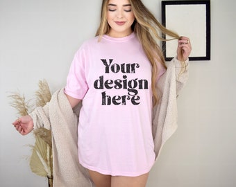 Pode incluir: Camiseta rosa claro com o texto "Your design here" em preto, com um efeito desgastado. Um cardigan bege está drapeado sobre os ombros. Uma imagem emoldurada está pendurada na parede ao fundo.