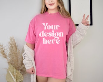Pode incluir: Uma t-shirt rosa vibrante com a frase branca "Your design here". É usada com um cardigan de cor clara. A imagem sugere um estilo descontraído e confortável, adequado para o uso diário.
