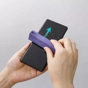以下が含まれることがあります： 黒いスマートフォンを手に持ち、紫色のマイクロファイバークロスで画面を拭いている。画面は濡れていて、水滴がついている。