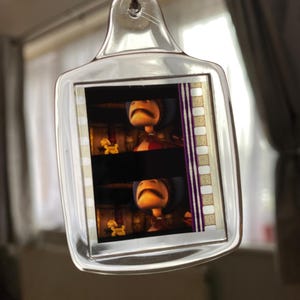 May include: A clear plastic keychain with a film strip design featuring two images of a cartoon character with a surprised expression. The character is wearing a red shirt and a brown hat.