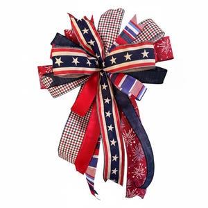 May include: A decorative bow featuring patriotic colors and patterns. The bow includes red, white, and blue ribbons with star and stripe designs, as well as a checkered pattern. The bow is suitable for various decorative purposes.