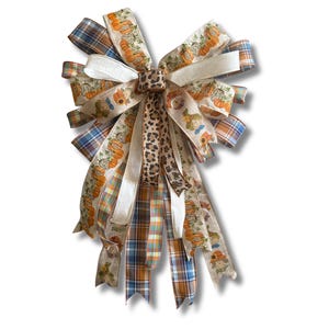 May include: A decorative bow made of multiple ribbons featuring a mix of patterns including orange pumpkins, sunflowers, scarecrows, blue and orange plaid, solid cream, and a central leopard print strip with long trailing ribbon tails.