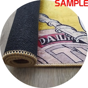 Puede incluir: Alfombra enrollada con una parte inferior negra y texturizada y un borde beige. La parte superior de la alfombra presenta un dise&ntilde;o de dibujos animados con la palabra "DAILY" en una pancarta roja. La palabra "SAMPLE" est&aacute; en rojo en la esquina superior derecha.