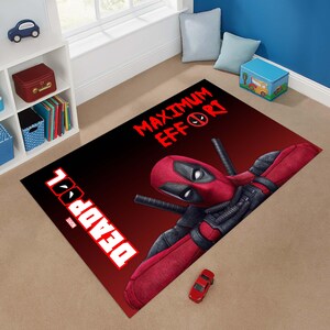 May include: A rectangular rug featuring the Deadpool character in a red and black color scheme. The rug has the words "DEADPOOL" and "MAXIMUM EFFORT" in white and red text. A red toy car is on the rug.