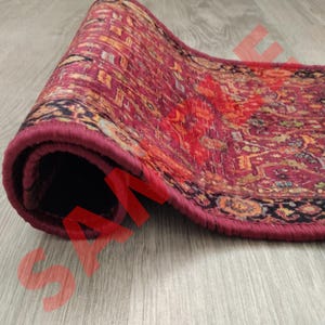 Pu&ograve; includere: Un tappeto da corridore arrotolato con un motivo rosso, arancione e nero e un bordo marrone scuro. Il tappeto &egrave; arrotolato e il bordo del tappeto &egrave; visibile.