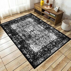 Könnte beinhalten: Ein rechteckiger Teppich mit einem verzierten, traditionellen Design. Der Teppich hat ein zentrales Feld in Hellgrau und Weiß, das von einem schwarzen Rand mit Blumen- und Scrollmustern umrahmt wird. Der Teppich liegt auf einem Holzboden.