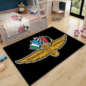 May include: Black area rug with a gold Indianapolis Motor Speedway logo. The logo features a winged circle with the words "Indianapolis Motor Speedway" and "Motor" inside the circle. The wings are spread out and have a checkered flag design on the back.