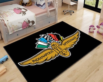 May include: Black area rug with a gold Indianapolis Motor Speedway logo. The logo features a winged circle with the words "Indianapolis Motor Speedway" and "Motor" inside the circle. The wings are spread out and have a checkered flag design on the back.