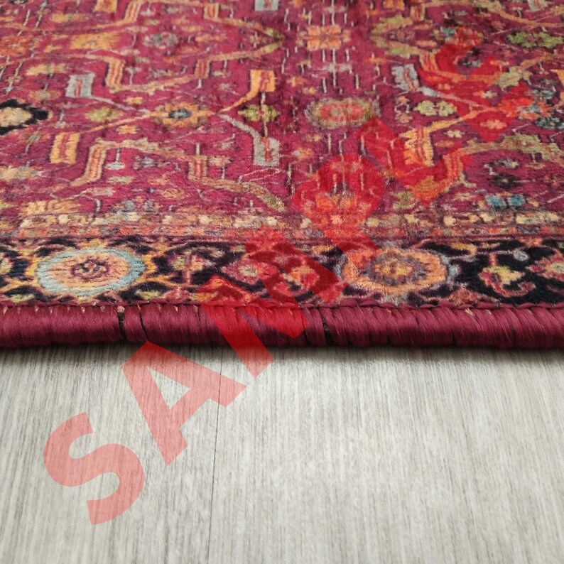 Pu&ograve; includere: Primo piano del bordo di un tappeto rosso, arancione e blu con un bordo marrone scuro e frange. Il tappeto &egrave; su un pavimento in legno chiaro.