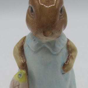 May include: A ceramic rabbit figurine, brown with a light blue dress and a basket. The rabbit stands on a green base. The figurine has detailed features and a classic design, reminiscent of a children's story.