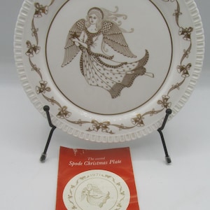 May include: A white Spode Christmas plate from 1971, with a brown angel design. The plate has a decorative border with bells and bows. It's displayed on a black metal stand, with a red booklet that reads "The second Spode Christmas Plate".