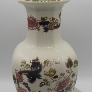 May include: Cream ceramic table lamp with floral and gold detailing. The lamp has a vase shape, a white top, and a dark brown base. The floral design features pink, blue, and green elements. The lamp is plugged in.