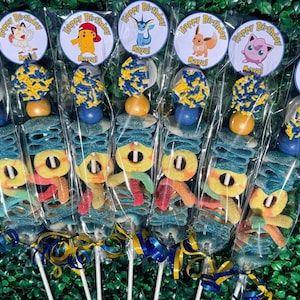 May include: Candy skewers featuring colorful gummy candies and lollipops. Each skewer is individually wrapped and topped with a circular sticker that says "Happy Birthday" and a cartoon character. The skewers are decorated with blue and yellow ribbons.