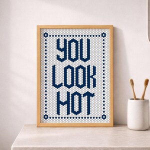 May include: A framed print with the words "YOU LOOK HOT" in navy blue block letters. The text is set against a white honeycomb pattern, framed by a light wood border. The print is on a bathroom counter.