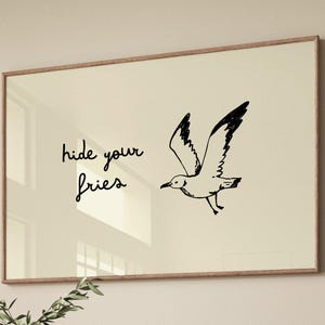 May include: A framed print featuring a beige background with a black illustration of a seagull in flight. The text "hide your fries" is written in a cursive font. The frame is a light brown color.