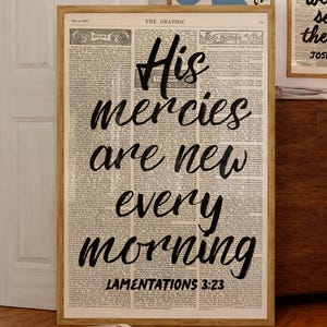 Könnte beinhalten: Ein gerahmter vertikaler Wandkunstdruck mit dem Text His mercies are new every morning LAMENTATIONS 3:23 in schwarzer Schreibschrift auf einem Hintergrund aus einer alten Zeitung. Der Druck steht auf einem Holzboden neben einem Schrank und zwei weiteren Bildern.