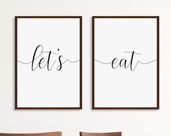 May include: Two framed prints with the text "let's" and "eat" in a cursive font. The prints are hanging on a white wall above a wooden table with chairs.