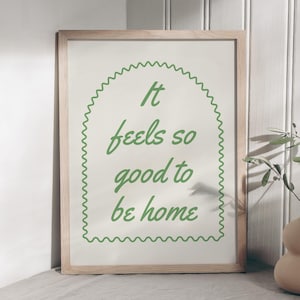 Może przedstawiać: Oprawiony wydruk z napisem "It feels so good to be home" w zielonym skrypcie. Wydruk jest w jasnej drewnianej ramie i leży na szarej powierzchni. Po prawej stronie znajduje się beżowy wazon z zielenią.
