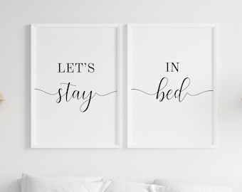 May include: Two framed art prints with the words "LET'S stay" and "IN bed" in black script on a white background. The prints are displayed above a bed with white pillows and a beige blanket. The overall theme is bedroom decor.