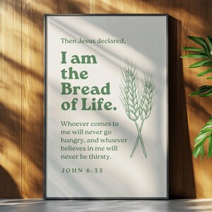 Op de afbeelding: Een ingelijste print met de tekst "Ik ben het Brood des Levens" in groene letters, vergezeld van een illustratie van tarwearen. De print wordt gepresenteerd tegen een houten muur, met een plant in pot op de achtergrond.