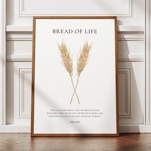 May include: A framed print featuring the text "BREAD OF LIFE" and a Bible verse from John 6:35. Two wheat stalks are crossed in the center. The frame is brown. The print is displayed against a white wall.