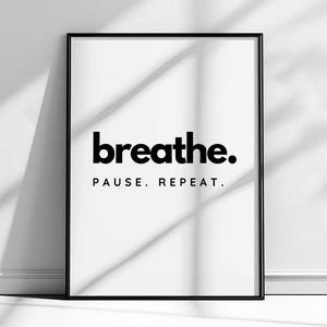 May include: A black framed print with the words "breathe. PAUSE. REPEAT." in bold black font on a white background. The frame is black. The print is leaning against a white wall, with shadows cast across it.