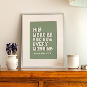 May include: A framed print with a sage green background and white text that reads "His mercies are new every morning" with the scripture reference "Lamentations 3:22-23" below. The print is sitting on a wooden table with a vase of lavender, a candle, and a lamp.