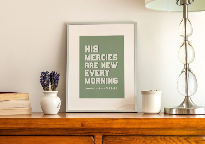 May include: A framed print with a sage green background and white text that reads "His mercies are new every morning" with the scripture reference "Lamentations 3:22-23" below. The print is sitting on a wooden table with a vase of lavender, a candle, and a lamp.