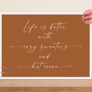 May include: A framed print with a white border and a warm brown background. The white cursive text reads "Life is better with cozy sweaters and hot cocoa." A pink "Digital Download" sticker is in the upper right corner.