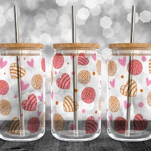 Puede incluir: Tres vasos de vidrio transparentes con tapas y pajitas. Cada vaso tiene un fondo blanco con un patrón repetido de pasteles en forma de corazón rosa y marrón.
