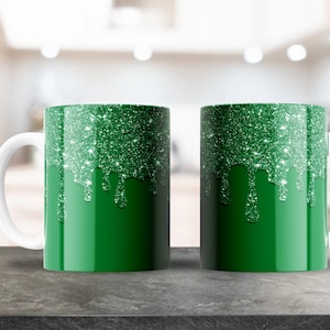 May include: Two white ceramic mugs with green glitter dripping down the sides. The mugs are on a gray countertop.